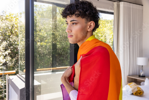 Man standing by sliding glass door to balcony with rainbow pride flag over shoulders, copy space