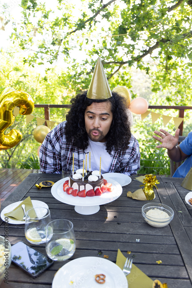 Obraz premium Group of friends gathering on wooden deck, hispanic male blowing out cake candles in party hat