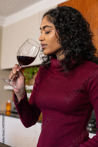 Indian woman sniffing red wine in glass near kitchen counter with herbs and orange bottle
