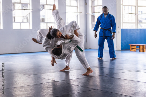 Diverse judo class practicing throws in white, blue gis on dojo mats near windows