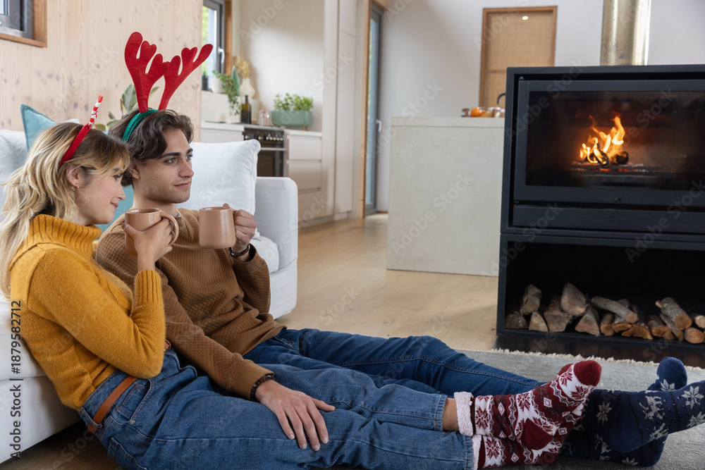 Naklejka premium Couple holding ceramic mugs and wearing antler headbands sitting by fireplace in living room