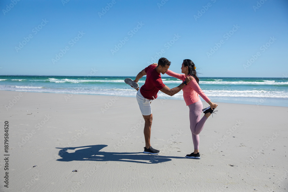 Fototapeta premium Diverse couple performing quad stretch on sandy beach by sea waves in sportswear and sneakers