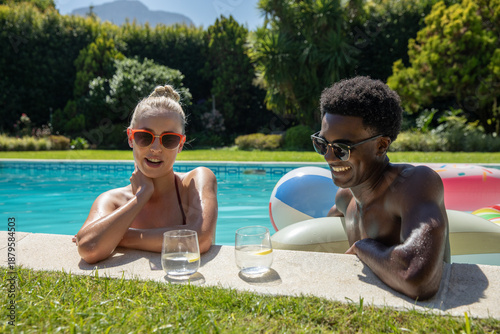 Diverse couple leaning on edge at home pool in swimwear holding pool tube, lemon-slice glasses