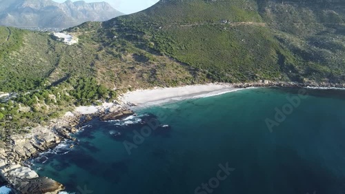 Aerial drone video of Sandy Bay beach coastline in Cape Town South Africa; top view of white sand beach with granite boulders and green mountainside on the Atlantic Ocean coast.