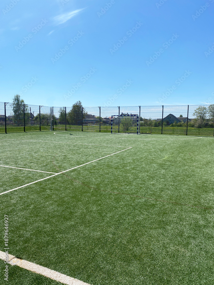 Obraz premium Green artificial turf sports field with goalposts and surrounding fence under a clear blue sky, ideal for mini-football or handball activities with ample space for play