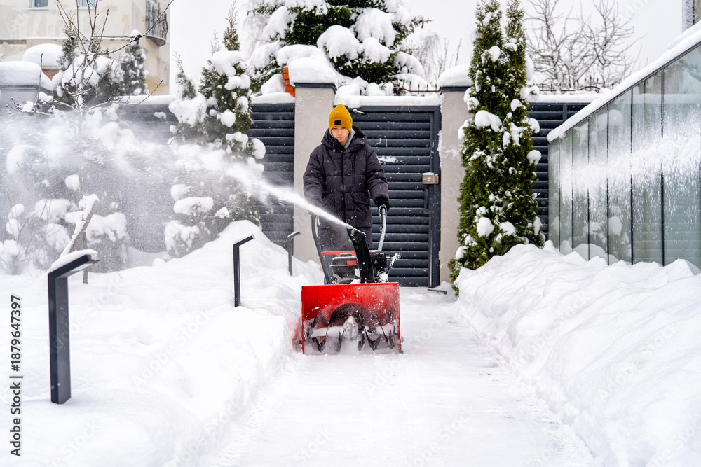 Naklejka premium Person diligently operates powerful red snow blower, efficiently clearing snow-covered pathway in front of modern house amidst winter wonderland scene, managing heavy snowfall.