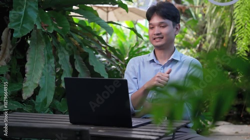 Young Asian businessman having a video meeting on his laptop while sitting in a lush green garden