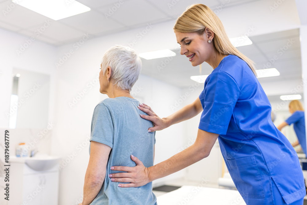 Obraz premium Female physical therapist in blue scrubs providing postural correction and back support to an elderly woman in a rehabilitation center, helping with spinal alignment and geriatric healthcare.