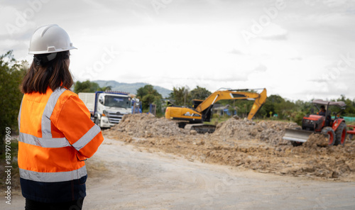 Photos A person wearing a hard hat and safety jacket stands by the road watching workers perform tasks with heavy machines