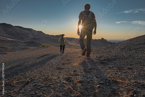 Rear view of two hikers, a man and a woman, walking on a dusty desert trail at sunset, with the sun low on the horizon creating silhouettes and a dramatic lens flare.