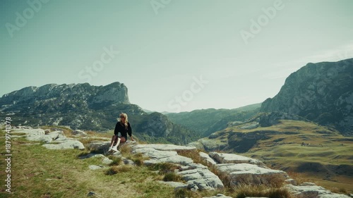 A woman in sunglasses sits on white limestone slabs at a grassy plateau as peaks and a tree lined valley frame a slow steady pan and a pale sky with crisp rock detail.