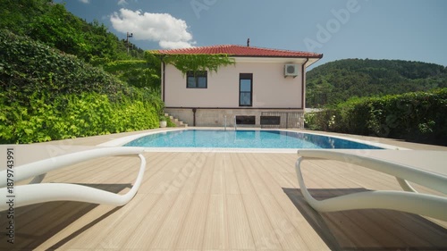 Low angle view of a private villa, red tiled roof, AC unit, blue mosaic pool and white lounge chairs as the camera dollies forward, wide and sunlit at midday