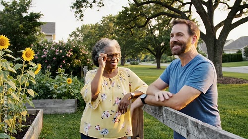 A cheerful mature African American woman points at blooming sunflowers in a raised garden bed while engaging in a friendly, smiling conversation with a middle aged Caucasian man leaning on a rustic.