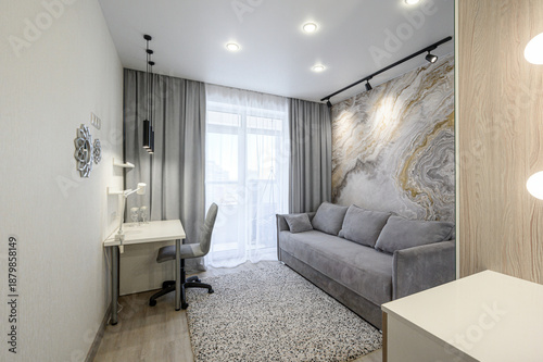 modern narrow living room with a grey sofa, a study area with a white desk and chair, marble-patterned accent wall, light grey curtains, a textured rug, and recessed ceiling lighting
