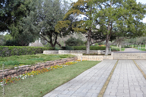 Beautifully manicured gardens at Ramat Hanadiv Nature Park in Israel. Featuring limestone retaining walls, lush green lawns, blooming flower beds, and stone paved walkways under Mediterranean trees. 