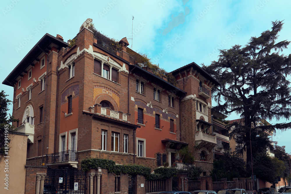 Obraz premium Fairy-tale villa in Quartiere Coppedè, Rome, by Gino Coppedè, with mosaic-tiled facade, arched entrance, tower with clock, balconies, and ornate details in warm tones under cloudy sky.
