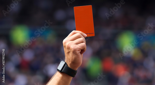 A close-up photograph of a caucasian male hand wearing a black smart watch holding up a bright orange-red rectangular card against a blurred stadium background.