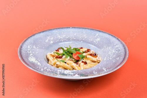 Creamy penne pasta with bacon and arugula served in a ceramic bowl isolated on orange background