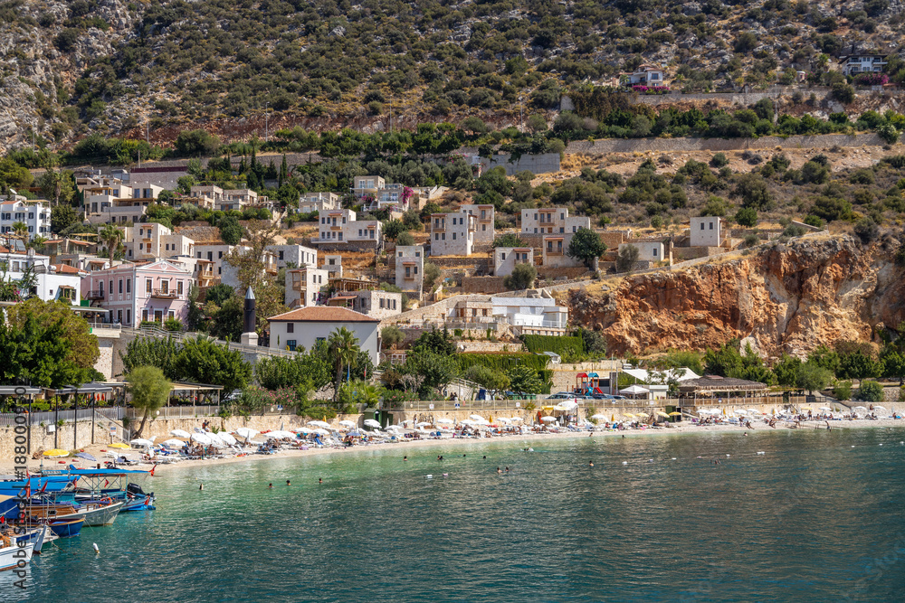 Fototapeta premium Scenic view of Kalkan town beach with white umbrellas and turquoise water. Mediterranean coastal landscape with traditional houses on green hills, shot from marina pier. September, Turkey.