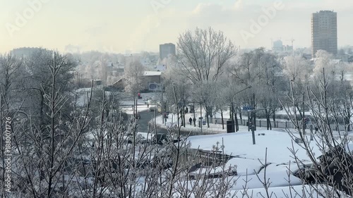 High-angle view of snow-covered city street, frost-covered bare trees in foreground. Urban life with moving cars on road and pedestrians on sidewalks.