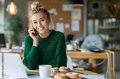 Smiling young woman talking on smartphone at office workspace with coffee and donuts