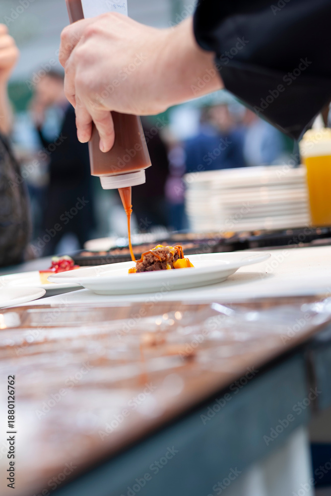 Fototapeta premium Chef drizzling sauce onto plated dish at culinary event or restaurant. Professional food preparation and fine dining presentation for hospitality, catering, and food service content.