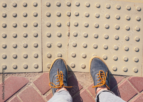 Women's feet in sneakers stand on a tactile tile. The road for the blind.