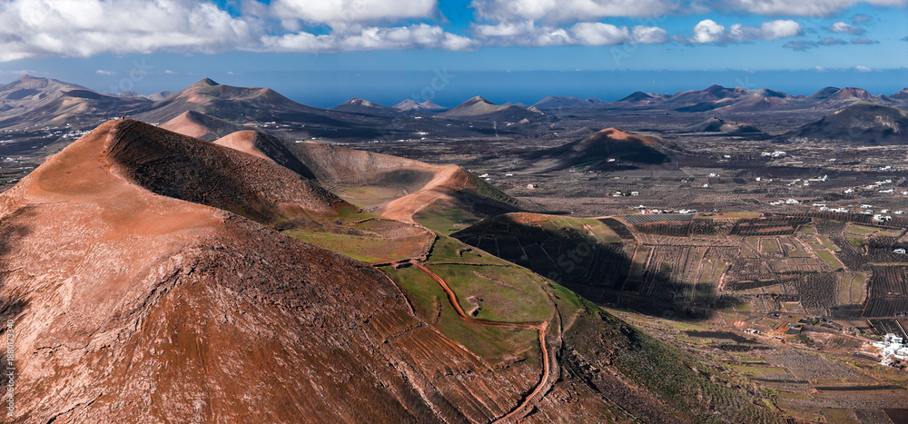 Fototapeta premium Aerial view shows Lanzarote, Canary Islands, with ochre caldera ridges, La Geria vine pits and stone walls, white houses, and conical volcanoes under patchy daylight.