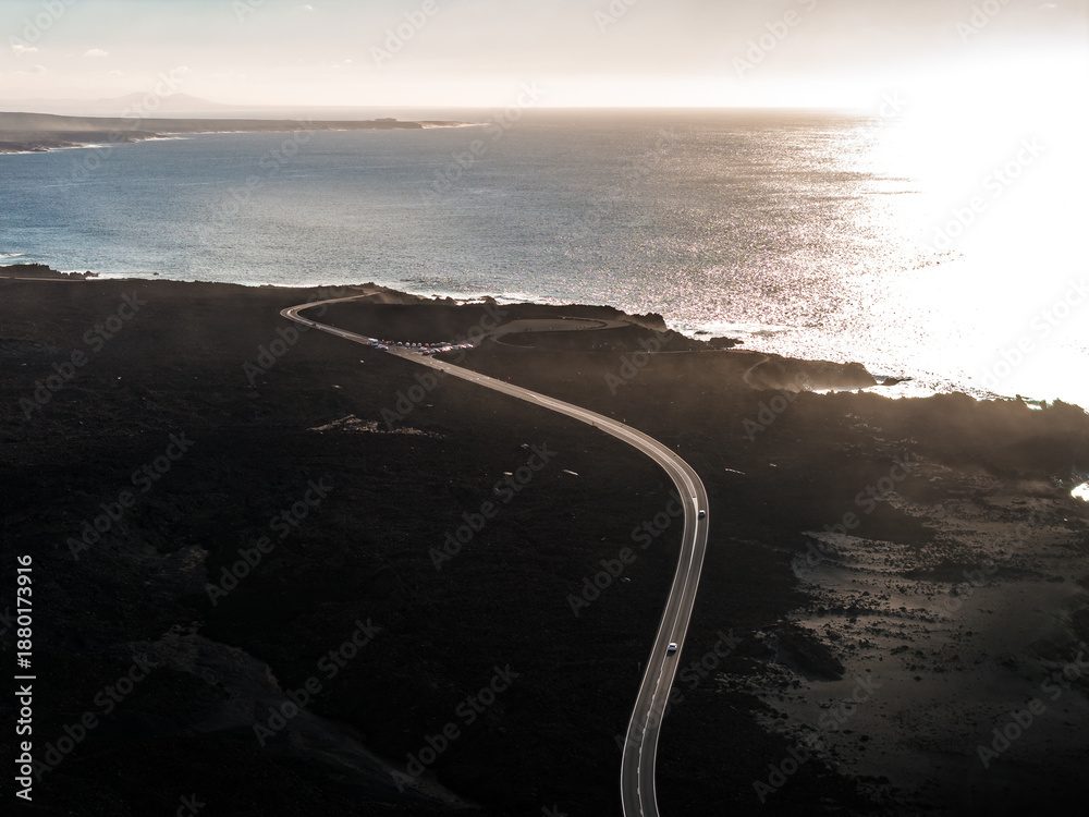 Fototapeta premium Aerial view shows a curving coastal road over black lava in Lanzarote, Canary Islands, near Timanfaya and El Golfo, with few cars and late afternoon light on the sea.
