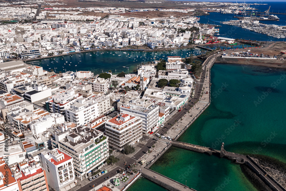 Obraz premium Aerial view of Arrecife, Lanzarote shows Charco de San Gines with small boats, a promenade, the church of San Gines, a stone causeway, breakwater, and the working harbor.