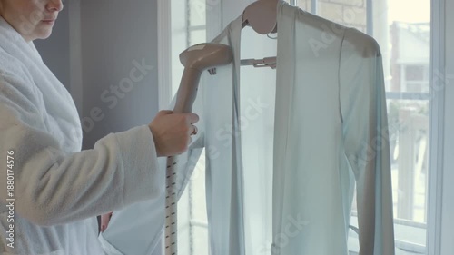 Anonymous woman in robe steaming a silk cardigan with a steamer in a cozy bedroom. The models face is not visible. Focus on hands and action.