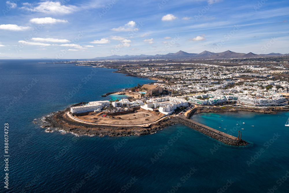 Fototapeta premium Aerial view of Costa Teguise, Lanzarote, shows curved breakwater, rocky shore, turquoise coves, hotel complexes, and a sandy construction area under clear daylight skies.