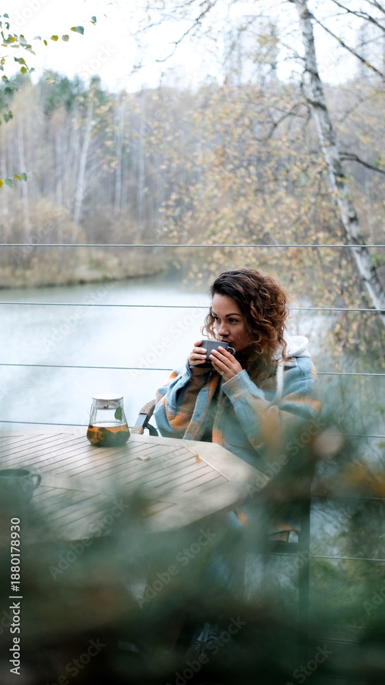 Naklejka premium A sweet woman with curly hair drinks tea and relaxes on the terrace of her house on an autumn day. Health care, sincerity, a sense of balance and calm.