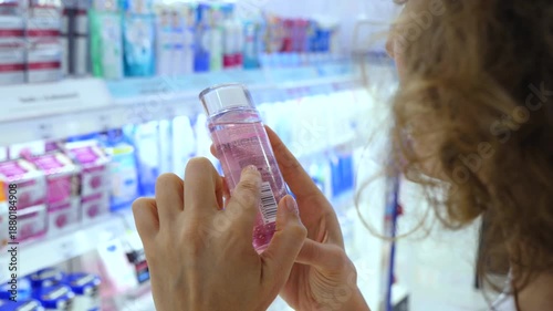 Close up of a young woman's hands holding a bottle of pink micellar water or toner, carefully reading the ingredient list and product information on the label in a beauty store aisle