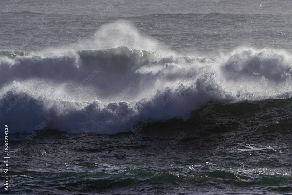 Naklejka premium A dark blue colored rip curl of a wave crashing on shore. The dangerous ocean swell has white foam and a water spray. There's a break in the center of the stormy surf with foam on both sides.