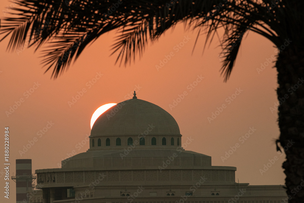 Obraz premium The sun sets behind a dome structure while a palm tree stands in the foreground. The scene captures the end of the day with warm colors in the sky.