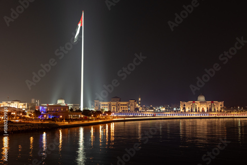 A tall flag pole is lit up at night, showing a large flag. Buildings and lights reflect in the water. People walk along the waterfront enjoying the evening scene.