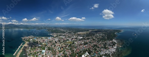 Wide-angle aerial panorama of Puerto Princesa City showcasing urban sprawl, airport runway, coastal waters, and mountain ranges under blue sky in Palawan, Philippines.