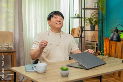 Obraz na płótnie Asian man sitting at home table finishes work, closes laptop, removes glasses, rests back in chair with happy sigh