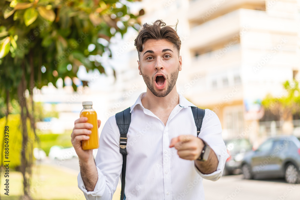 Fototapeta premium Young handsome man holding an orange juice at outdoors surprised and pointing front