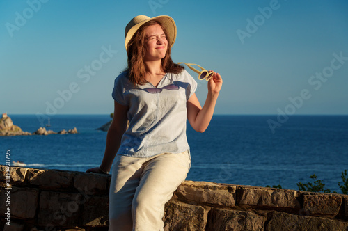 Foto Portrait of a young woman squinting at the bright sunset sun, holding her sunglasses and wearing a straw hat by the seaside during holidays