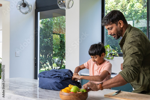 Indian father and son standing at marble island countertop unpacking blue backpack, copy space