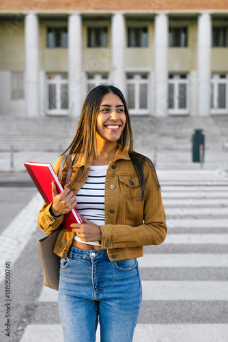 Young woman confidently walking on campus, holding red notebooks, representing higher education concepts like learning, success, and diversity
