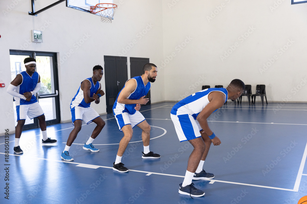 Fototapeta premium Diverse male teammates guarding in low stance on gym court with basketball hoop