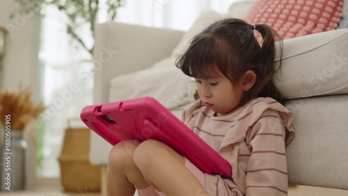 Asian young girl sitting at home and focusing on playing a game on a tablet, a female child enjoying calm and quiet digital screen time.