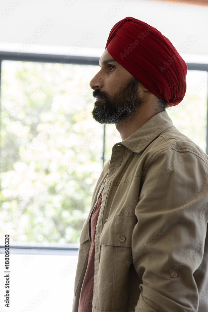 Obraz premium Indian man wearing red turban and jacket by window in natural light, gazing at foliage, copy space