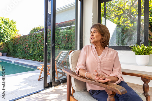 Senior woman sitting in wooden armchair by sliding glass door at home overlooking pool, copy space