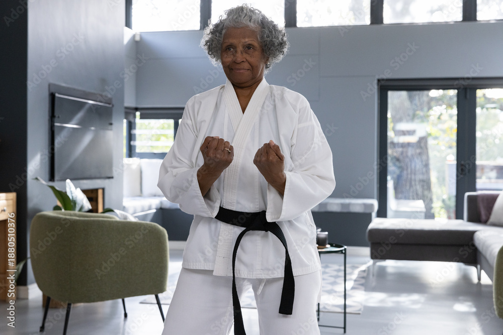 Fototapeta premium Senior african american woman holding fighting stance in living room wearing karate gi and belt