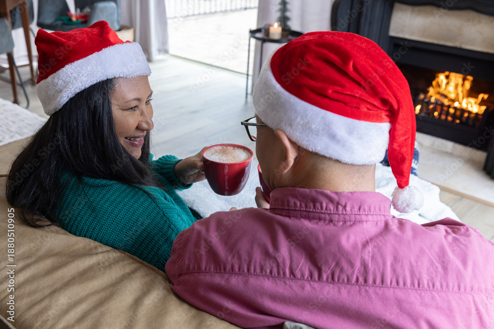 Fototapeta premium Diverse couple in santa hats sipping beverages from red mugs on beige couch beside fireplace