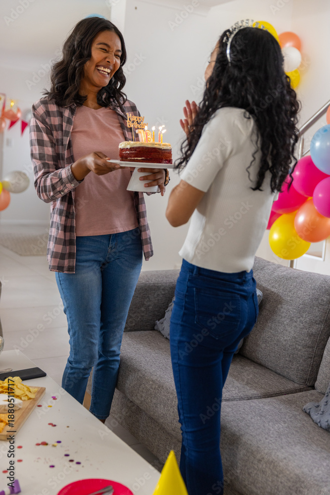 Obraz premium Diverse mom and teenage daughter holding birthday cake with lit candles at home near balloons